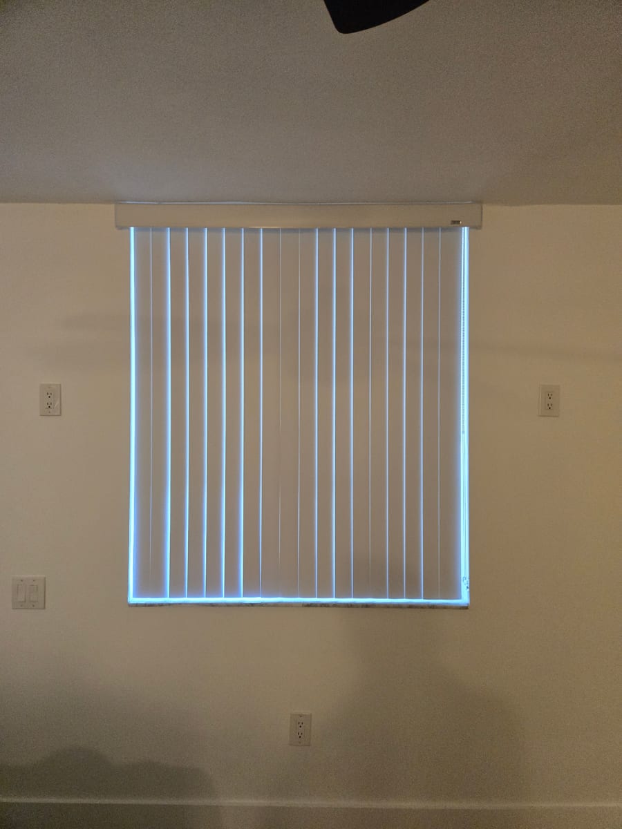 A bedroom window fitted with old white vertical blinds.